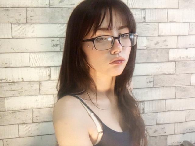 Eilene Szala, avec ses longs cheveux foncés, ses lunettes et son débardeur gris, qui révèle ses petites seins et affiche une expression neutre, se tient devant un mur de briques blanches..
