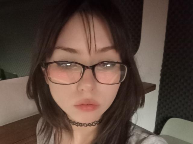 Eilene Szala, met bleke huid, donker steil haar, zwarte bril, roze blush op de wangen, kleine roze lippen, een zwarte choker, die recht in de camera kijkt, met een licht geopende mond..