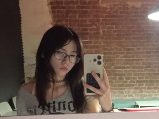 Eilene Szala, avec des lunettes, un collier noir et un haut gris à épaules dénudées, prend un selfie devant un miroir avec un iPhone blanc. Elle a de longs cheveux bruns et une expression sérieuse..
