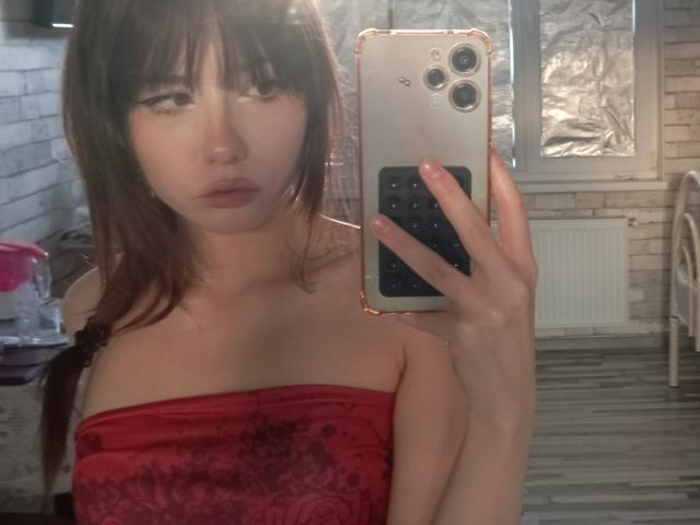 Eilene Szala, met bleke huid, bruin haar en een pony, draagt een rode top zonder bandjes en maakt een selfie in de spiegel met een iPhone, waarbij haar borsten zichtbaar zijn en ze een neutrale uitdrukking heeft..