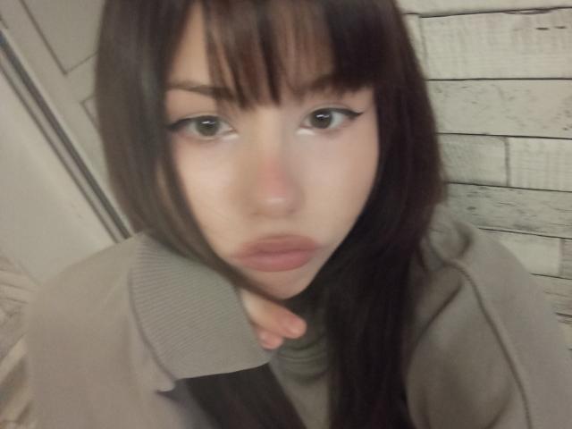 EileneSzala has brown eyes, full pink lips, and long dark brown hair with bangs, wearing a grey coat, looking into the camera with a slightly pouty expression.