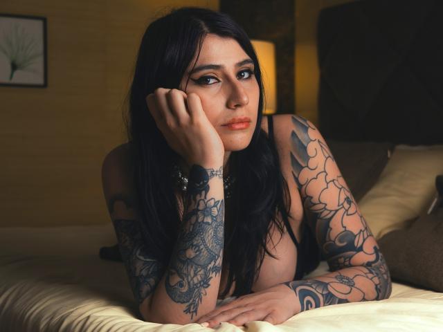 AlynaFerrer with long black hair, tattoos on both arms, resting her chin on her hand, lying on a bed, wearing a black bra, looking at the camera with a slight smile.