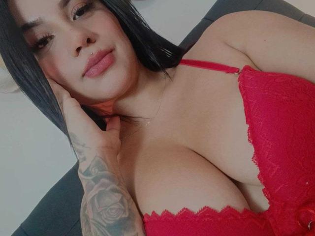 SamanthaLune with black hair, red lace bra, and tattooed hand, showing her big tits and nose ring, smiling softly.