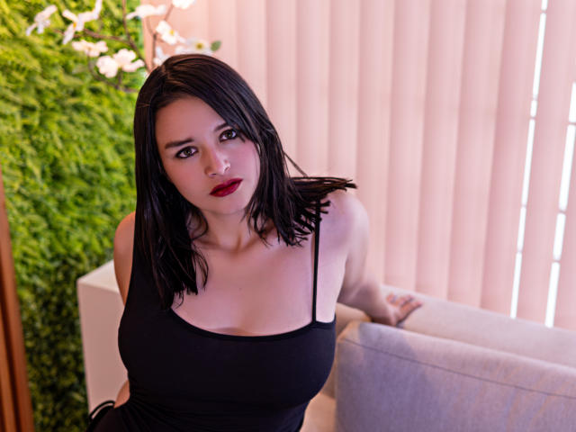LiaMoonroe, con i capelli neri, il rossetto rosso e una canotta nera che mette in mostra il seno, appoggiata a un divano grigio, guarda verso l'alto con un'espressione leggermente infastidita..