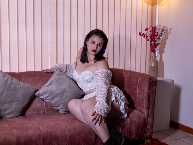 LiaMoonroe, avec une poitrine de taille moyenne, portant une robe blanche en dentelle à épaules dénudées, des bottes noires, assise sur un canapé rose, avec des cheveux foncés et mouillés, du rouge à lèvres rouge, des coussins gris, un vase avec des fleurs rouges..
