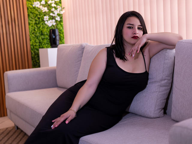LiaMoonroe, com cabelo preto, batom vermelho e vestido preto, reclinada em um sofá cinza, com um braço sobre uma almofada, olhando para a câmera, mostrando seus seios e nádegas..