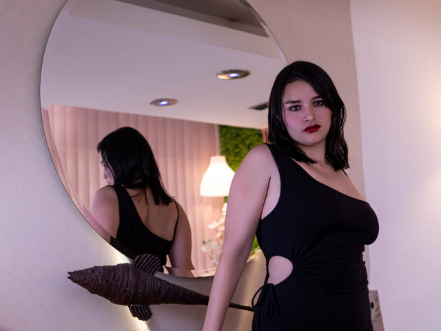 LiaMoonroe, con i capelli neri, il rossetto rosso e un seno abbondante, indossa un abito nero con aperture e sta in piedi di fronte a uno specchio, tenendo un cuscino marrone scuro..