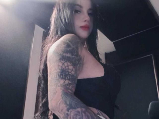 SamanthaLune has long black hair, red lipstick, and tattoos covering her right arm. She's wearing a black strapless top, showing her boobs. She's looking at the camera.