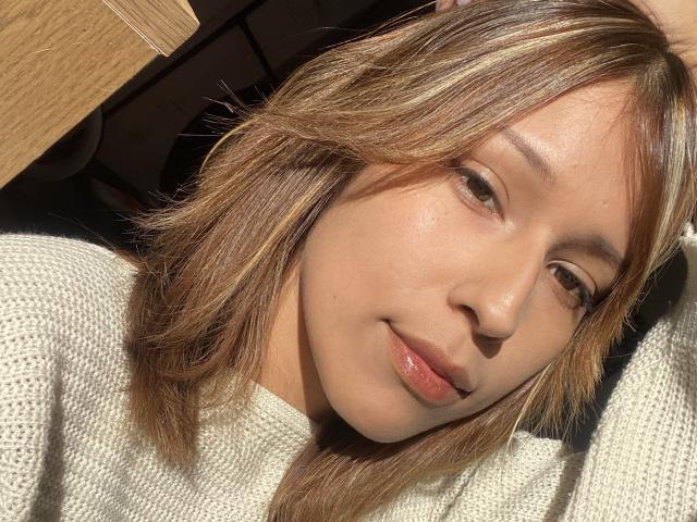 Il viso di KiaraHernandezX, con i capelli castano chiaro, un trucco leggero e un leggero sorriso, indossa un maglione beige in maglia, la luce del sole sulla pelle e una mano tra i capelli..
