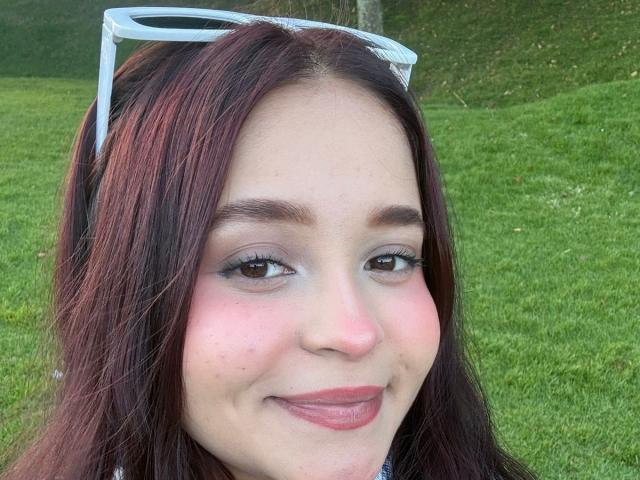 SamyMancini a la peau claire, les yeux marron et un rouge à lèvres rosâtre. Elle sourit, a les cheveux brun foncé, porte des lunettes de soleil blanches sur la tête et a une légère rougeur sur les joues..