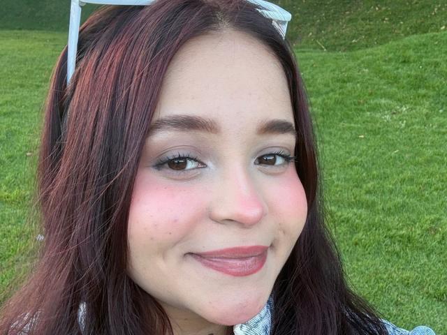 Das Gesicht von SamyMancini mit rosigen Wangen, braunen Augen und dunkelrotem Haar, sie trägt weiße Sonnenbrillen auf dem Kopf und lächelt mit pinkfarbener Lippenstift. Ihre Brüste sind nicht sichtbar..