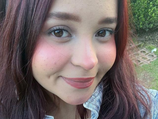 SamyMancini has a cute face with red-pink lipstick, brown eyes, and red-brown wavy hair. Her slightly flushed cheeks and subtle smile look sweet. She's wearing a gray shirt.