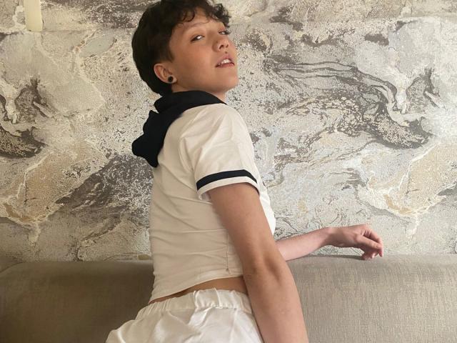 LissyLuxe with short brown hair, small earrings, white sailor shirt, and white pants, sitting on a grey sofa, looking over her shoulder, showing a slight butt and small chest.