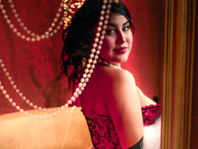AnnyReese with dark wavy hair, red lace corset, pearl necklace, and black gloves, looking back, pearls in front, smiling, red and gold lighting. Her tits are visible.