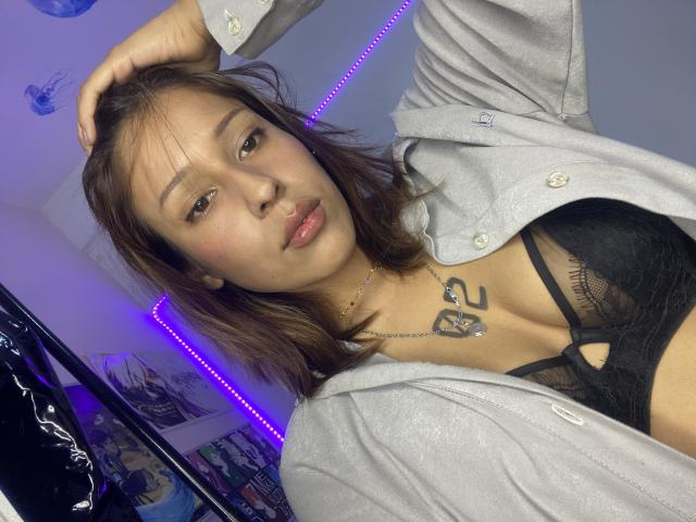 KiaraHernandezX, con capelli castani, pelle chiara e un tatuaggio sopra i suoi seni neri coperti da un reggiseno, indossa una camicia grigia sbottonata, con una mano tra i capelli, e una collana, in una stanza..