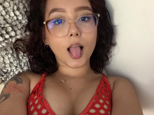 SophiaBrowm with curly dark hair, red glasses, and tongue out, wearing a red crochet top, showing her tits, and a flower tattoo on her right arm.