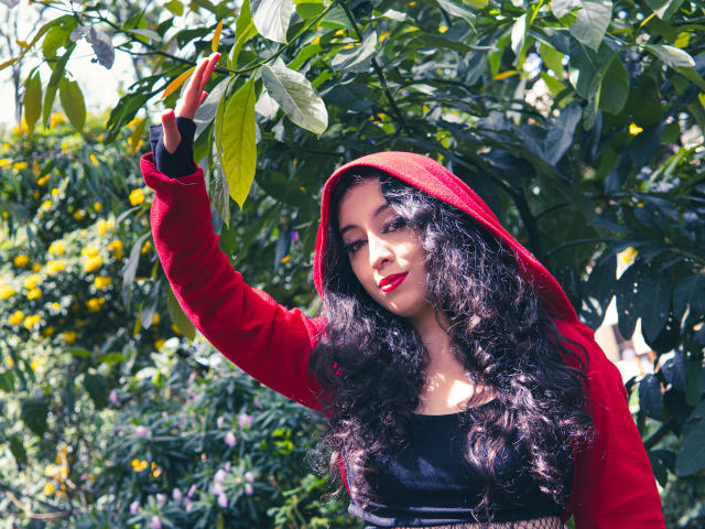 KendraSimons with long curly black hair, red hoodie, and black top, raising her right hand to green leaves, red lipstick, and confident smile in a sunny garden.