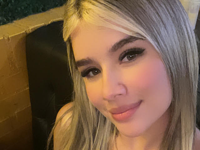 SashaPolet a les cheveux blonds, des sourcils épais, de longs cils noirs, un blush rose et un léger sourire. Ses lèvres roses sont légèrement pincées. Elle regarde directement l'appareil photo..