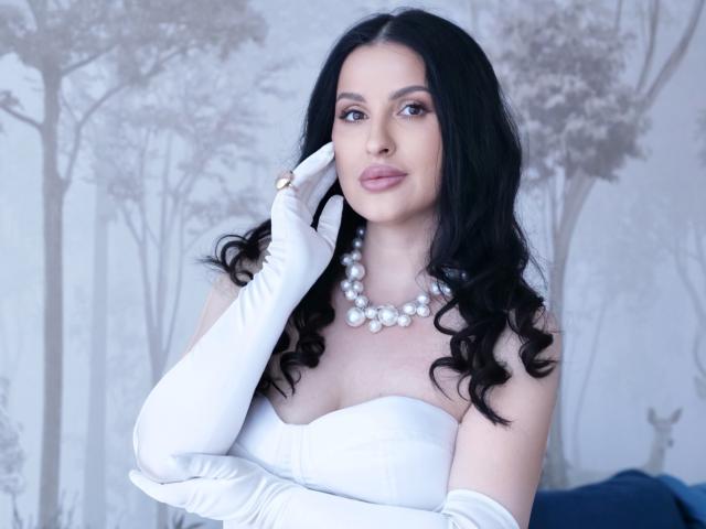 HoneyBlossom with black wavy hair, white strapless dress, pearl necklace, and white gloves, touching her face, showing her tits, with a blue upholstered seat in the background.