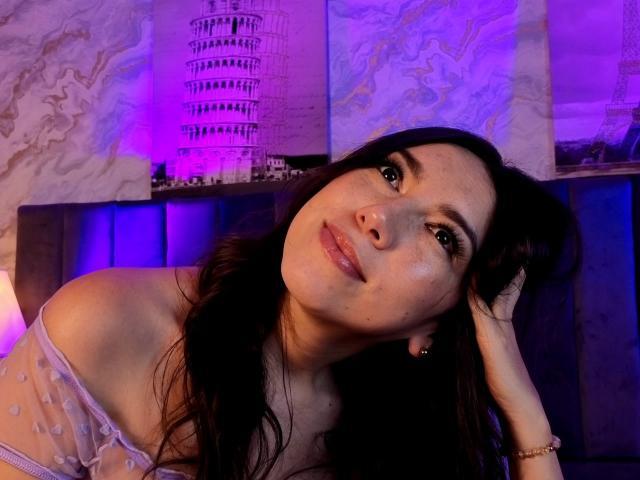 MiaReed with long dark hair, off-shoulder sheer top, looking at camera with a slight smile, purple and blue lighting, hand in hair, wearing a bracelet.
