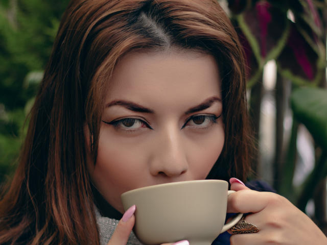 MandyMouses with brown hair, holding a white cup, has winged eyeliner, pink nails, and a ring, sipping, looking forward with focused eyes.