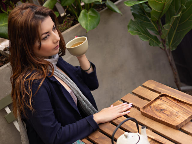 MandyMouses with brown hair, holding a beige cup, sitting at a wooden table, wearing a navy jacket and gray scarf, sipping, with a wooden tray and white teapot.