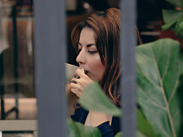 MandyMouses with brown hair, sipping from a cup, her eyes closed, wearing a dark shirt, holding the cup to her mouth, green plant leaves in the foreground.