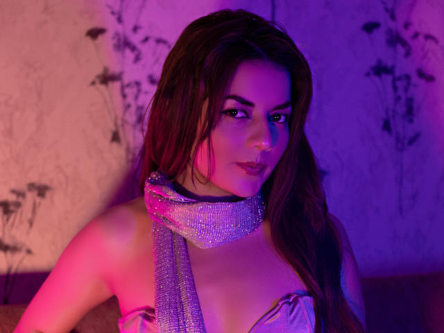 MandyMouses con lunghi capelli castani, che indossa una sciarpa argentata scintillante e una maglia viola illuminata con sfumature rosa, che mostra il seno, in una foto sexy e sensuale, illuminata di viola..