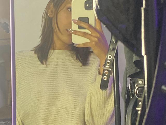 Kiara Hernandez, mit mittelbrauner Haut und glattem, braunem Haar, trägt einen beigen Strickpullover und macht ein Selfie vor einem Spiegel mit einem weißen Handy. Sie trägt ein schwarzes Lederarmband..