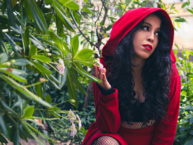 KendraSimons with curly black hair, red hoodie, and fishnet top, holding a green leaf, looking up with red lipstick, sitting in lush greenery, her tits partially visible.