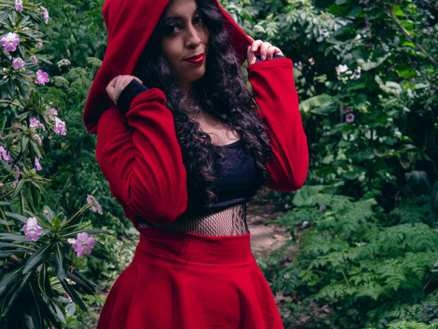 KendraSimons with long curly black hair, red hooded jacket, black mesh top, and red skirt, holding hood up, red lipstick, green forest, purple flowers, and a path.