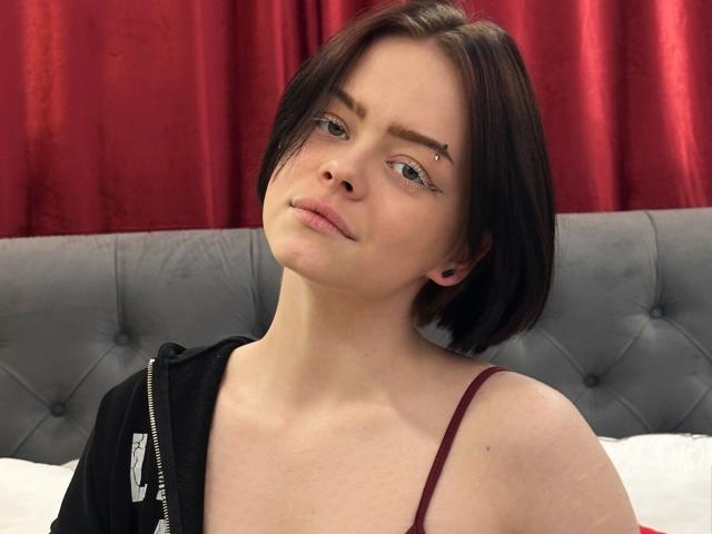 RiskyBeauty tiene el pelo corto y castaño, una sudadera con capucha negra y una correa color burdeos. Está mirando hacia arriba con una ligera sonrisa, y tiene un pequeño tatuaje de una estrella en la ceja derecha..