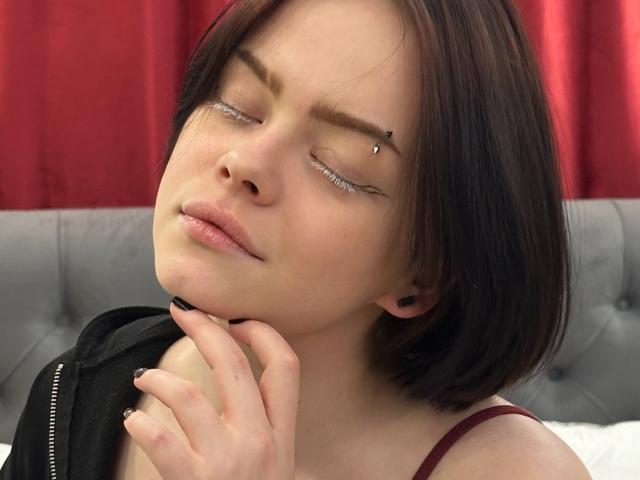 RiskyBeauty con capelli castani corti, occhi chiusi, anello al naso nero e piercing all'arcata sopraccigliare, che si tiene il mento, indossa una cinghia rossa, una giacca nera e tappi per le orecchie neri..