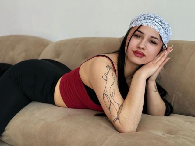 Dindinlir-hot liegt auf einem beigen Sofa, trägt ein rotes Tanktop und schwarze Hosen, und hat ein weißes Spitzen-Haarband. Ihr Hintern ist deutlich sichtbar, und sie hat ein Tattoo am Arm..