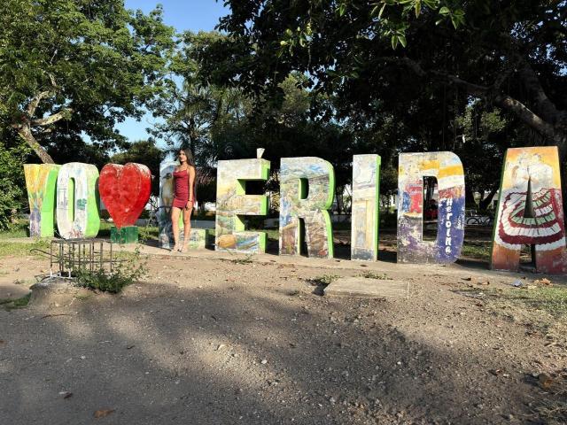 ScarlettRoss69 está atrás de letras coloridas e pintadas que formam a palavra "BEM-VINDO", com um coração vermelho e vários desenhos artísticos, em um parque ensolarado, cheio de árvores e com um caminho de terra..