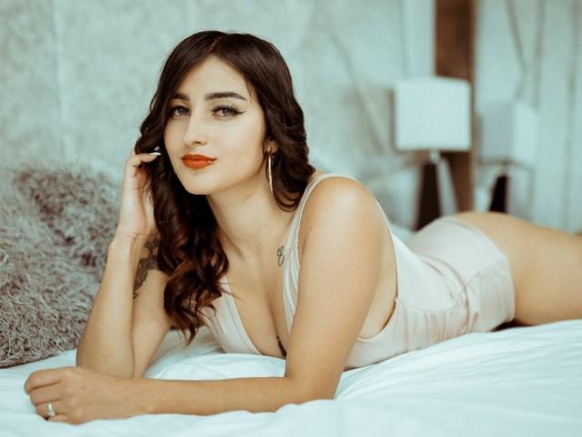 LisaNahyan with wavy brown hair, wearing a low-cut, beige one-piece, lying on a bed, looking at the camera with orange lipstick, touching her hair, showing her tits.