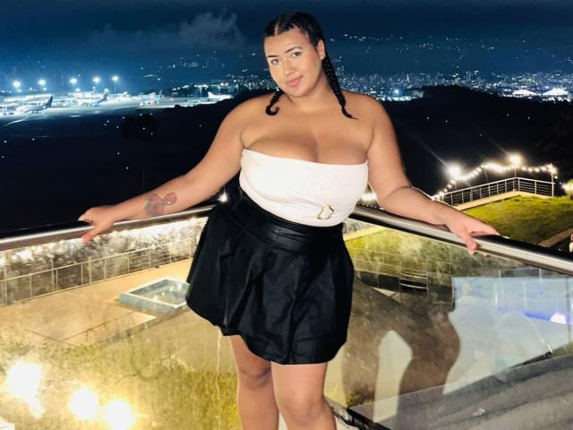 DojaAissha with big boobs in a white strapless top and black skirt, standing on a balcony at night, arms on the railing, tattoo on her left arm, looking cute.