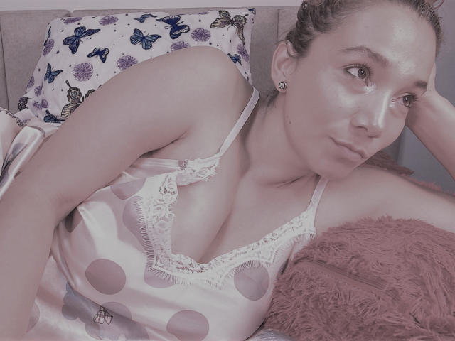 AmberDemons is lying on a bed, wearing a pink polka dot camisole with lace trim, showing her tits, with butterflies on a pillow and a pink fuzzy pillow.