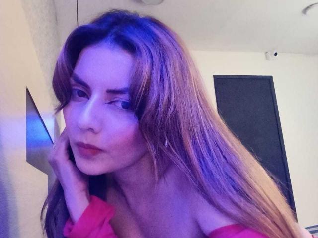 MandyMouses con lunghi capelli castani, occhi azzurri e labbra rosse, indossa una maglietta rosa con una spalla scoperta, mentre si tocca il viso in un selfie illuminato di blu. Il suo seno è leggermente visibile..