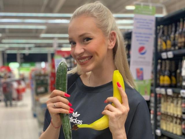 SylvieGray with blonde hair in a ponytail, holding a green cucumber and yellow banana, smiling, wearing a dark Adidas shirt, red nails, in a brightly lit grocery store.