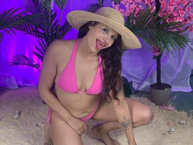 AriannaSeete, with pink bikini, straw hat, and tongue out, crouches on sand, showing her tits and pussy, tattoo on right arm, under purple light, surrounded by green plants.