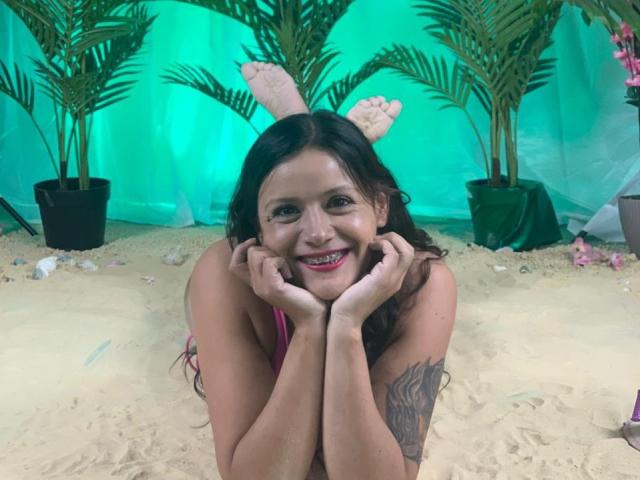 AriannaSeete, with long dark hair, pink bikini, and tan skin, smiles, hands near face, showing tattoo on right arm, with fake bunny ears and sandy beach background.
