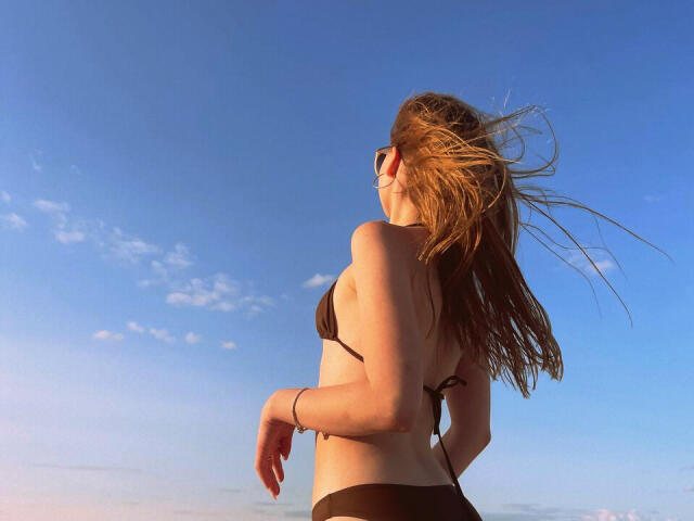 Nikollia with brown hair, wearing a black bikini, has her back to the camera, showing her small tits and bare ass, standing against a clear blue sky.