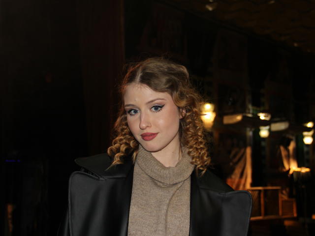 Nikollia, met krullend blond haar, blauwe ogen en rode lippenstift, draagt een zwarte leren jas en een beige coltrui, en heeft een schattelijk, sexy en aantrekkelijk gezicht..