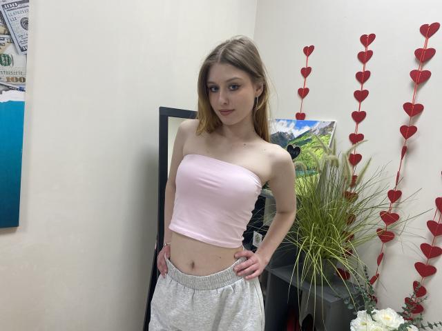 Nikollia, com seios pequenos, vestindo uma blusa rosa sem alças e calças de moletom cinzas, em pé com as mãos nos quadris, cabelo loiro, sorriso discreto, em um quarto com decorações de corações vermelhos..