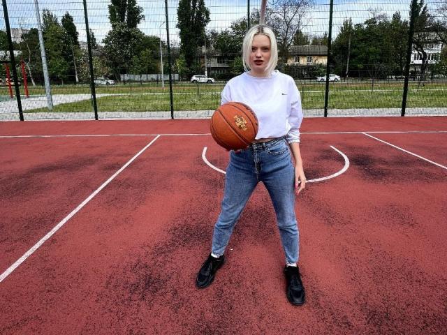 CrystalHope, com cabelo loiro curto, usando uma gola alta branca, jeans de cintura alta, botas pretas, segurando uma bola de basquete em uma quadra vermelha ao ar livre, em pé com as pernas afastadas, olhando para a câmera..
