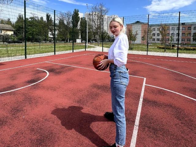 CrystalHope segurando uma bola de basquete, em pé em uma quadra vermelha ao ar livre, vestindo uma camisa branca, jeans azuis e tênis pretos, com o bumbum ligeiramente levantado e um sorriso adorável..