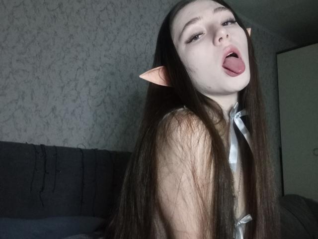 VeronicaAskwith with long brown hair, pale skin, and pink elf ears, tongue out, wearing a white necktie, looking up with a slightly open mouth.