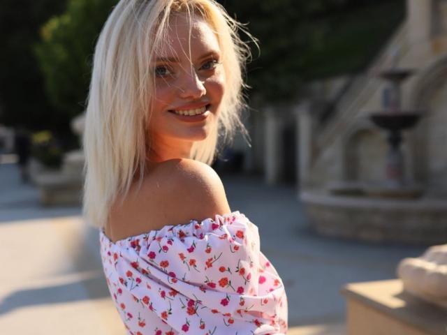 CrystalHope, com cabelo loiro, sorrindo, vestindo um vestido branco com um ombro de fora e estampas de cerejas vermelhas, iluminada pelo sol, olhando por cima do ombro, mostrando o ombro nu e um pouco de sua nádega..