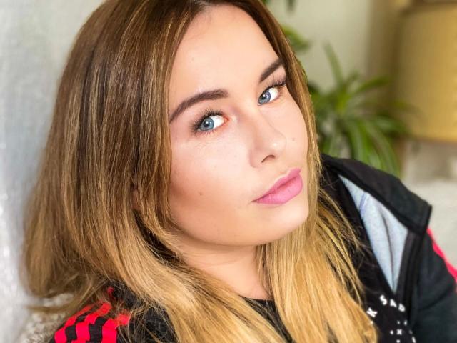 Shyla-hot ha i capelli biondi e lisci, gli occhi azzurri, il rossetto rosa e un naso leggermente all'insù. Indossa una giacca nera con strisce rosse. La sua espressione è neutra, e guarda verso la fotocamera..
